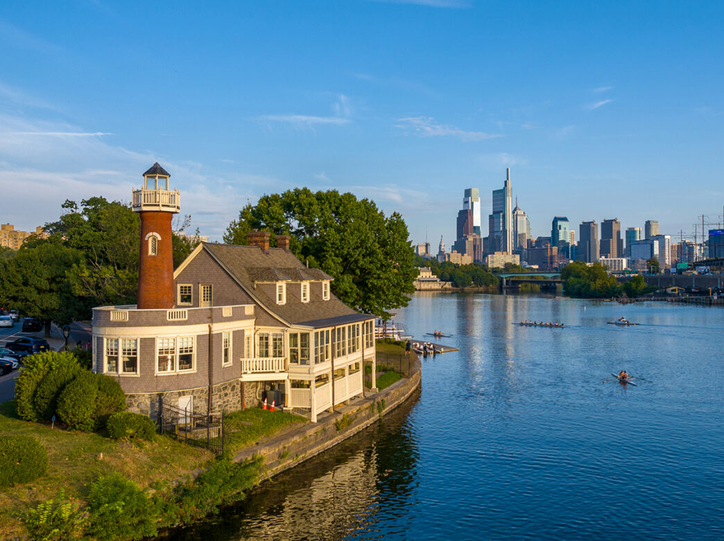 Un hangar à bateaux historique, surmonté d'une tour aux allures de phare, se dresse au bord de la rivière Schuylkill, tandis que des rameurs glissent sur l'eau, avec en arrière-plan la silhouette du centre-ville de Philadelphie se découpant sous un ciel bleu limpide.