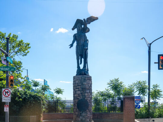 La estatua de bronce del jefe Tamanend se erige sobre un pedestal de piedra en la esquina de las calles Front y Market en Old City, con un águila posada sobre él y un cielo azul que enmarca el monumento.