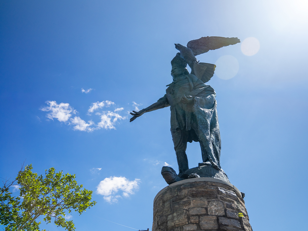 La statue en bronze du chef Tamanend trône au sommet d'un socle en pierre, surmontée d'un aigle perché au-dessus de sa tête, se dressant vers le ciel bleu éclatant parsemé de nuages épars.