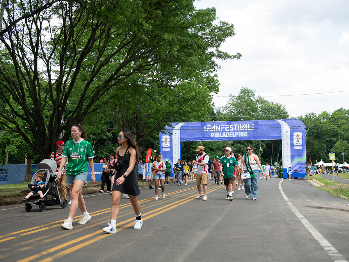 Visitors wearing soccer jerseys and team colors walk beneath the blue entrance arch to Philadelphia’s FIFA Fan Festival.