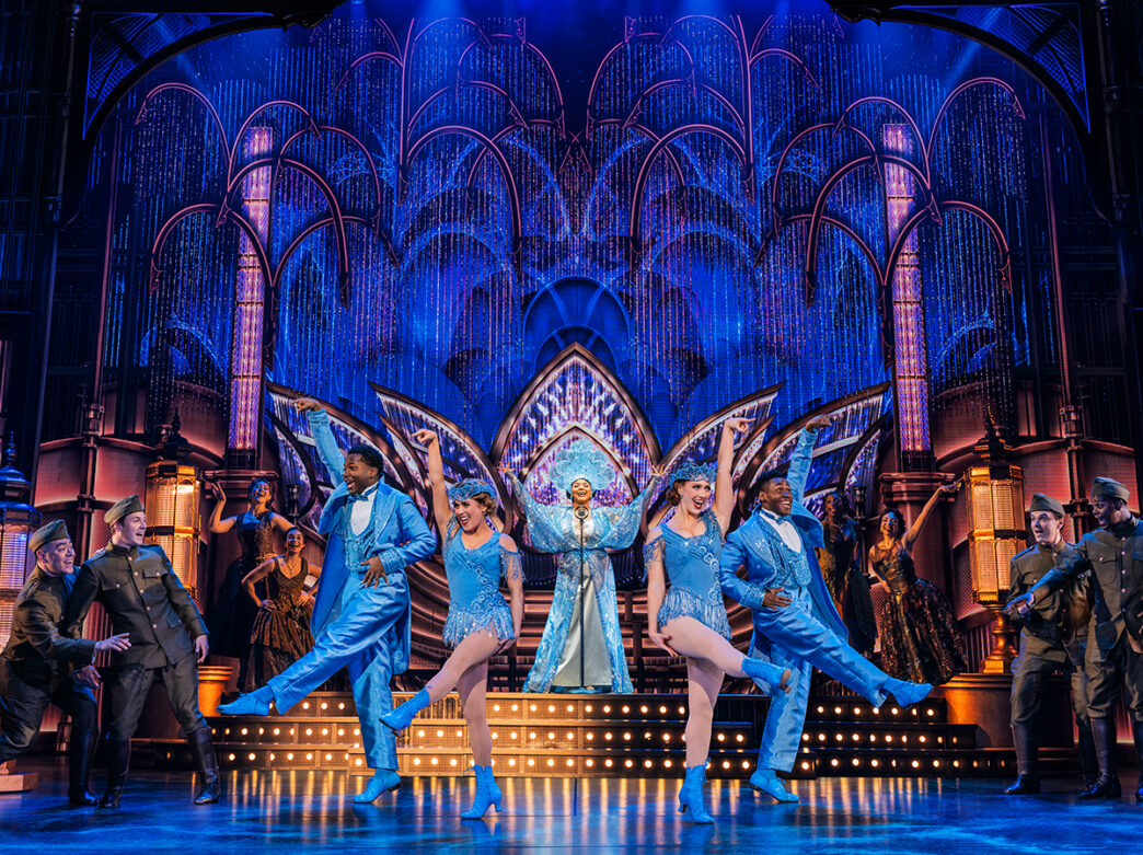 Performers in dazzling blue costumes sing and dance beneath an Art Deco-inspired set during a production of The Great Gatsby.
