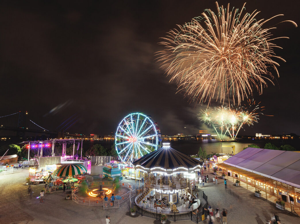 Golden fireworks explode over the colorful lights of Independence Blue Cross Riverrink Summerfest. A carousel, Ferris wheel, children's games and families can be scene in the forground.