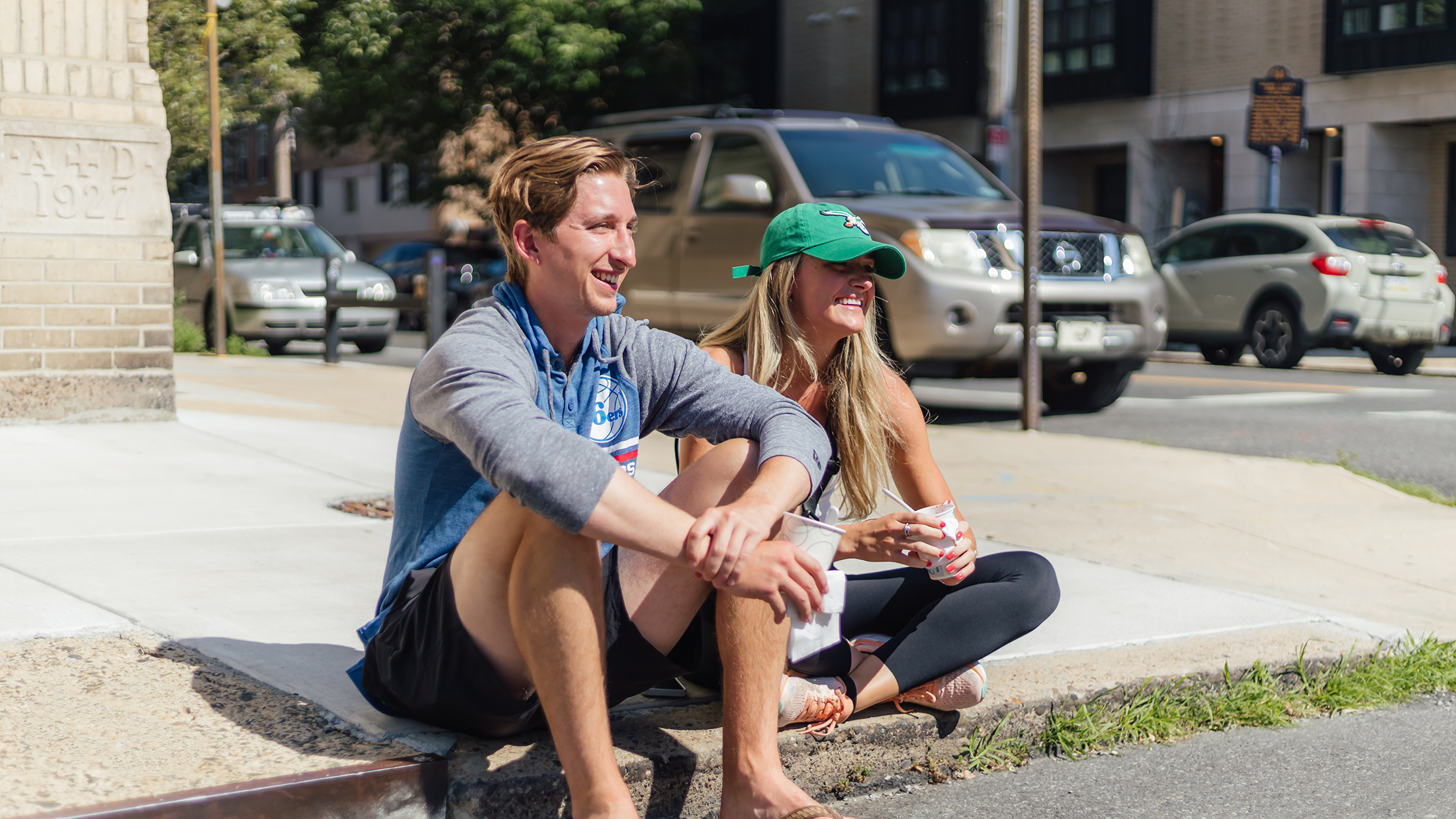 Deux personnes rayonnantes sont assises sur un trottoir ensoleillé devant le « John's Water Ice » ; elles sourient et savourent des coupes de glace à l'eau classique de Philadelphie par cette chaude journée.