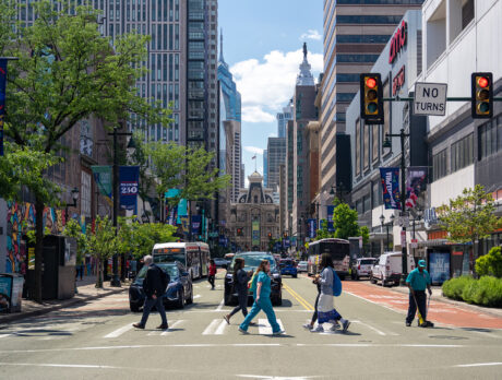 Os pedestres atravessam um cruzamento movimentado do centro da cidade ao longo da Market Street, cercados por arranha-céus, ônibus e faixas com temas da Filadélfia. O Philadelphia City Hall ergue-se ao longe, no final da rua, emoldurado por árvores e fachadas de lojas do centro da cidade.