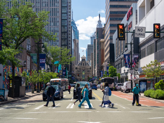 Los peatones cruzan una concurrida intersección del centro de la ciudad a lo largo de la calle Market, rodeados de rascacielos, autobuses y pancartas con motivos de Filadelfia. El edificio Philadelphia City Hall se alza a lo lejos, al final de la calle, enmarcado por árboles y las fachadas de las tiendas del centro.