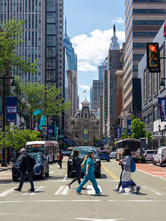 Pedestrians cross a busy Center City intersection along Market Street, surrounded by high-rise buildings, buses and Philadelphia-themed banners. Philadelphia City Hall rises in the distance at the end of the street, framed by trees and downtown storefronts.
