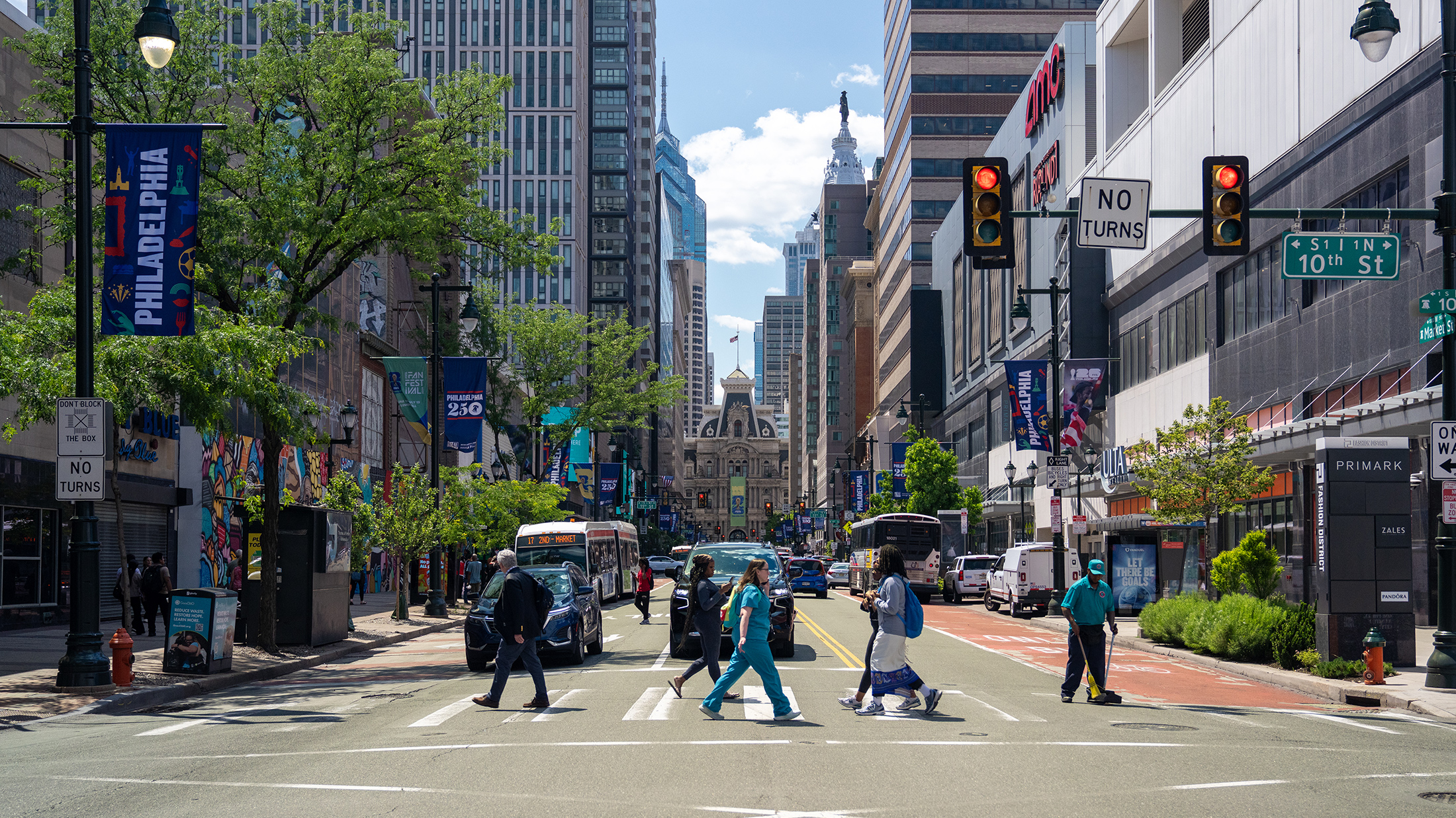 Voetgangers steken een druk kruispunt in Center City over langs Market Street, omringd door hoge gebouwen, bussen en spandoeken met motieven uit Philadelphia. In de verte, aan het einde van de straat, doemt Philadelphia City Hall op, omlijst door bomen en winkelpuien in het centrum.