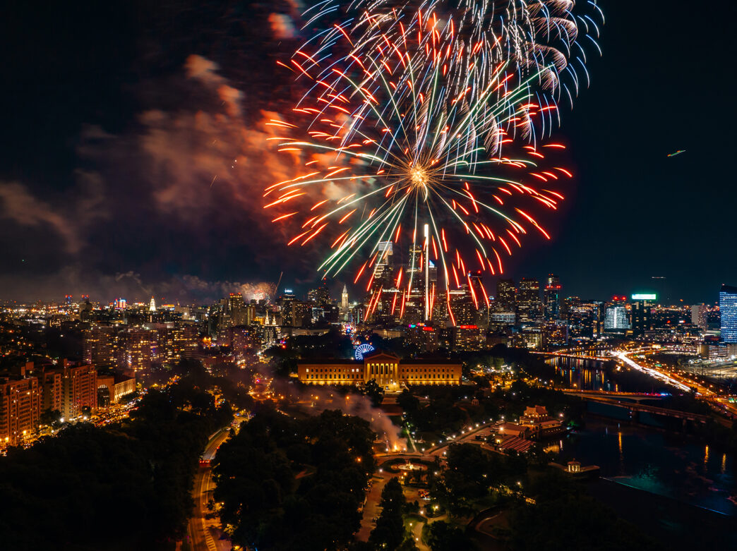 Fireworks burst above the Philadelphia skyline at night, illuminating the Benjamin Franklin Parkway, the Philadelphia Museum of Art and Center City skyscrapers during a Fourth of July celebration.