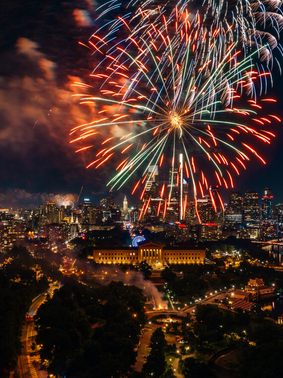 Fireworks burst above the Philadelphia skyline at night, illuminating the Benjamin Franklin Parkway, the Philadelphia Museum of Art and Center City skyscrapers during a Fourth of July celebration.