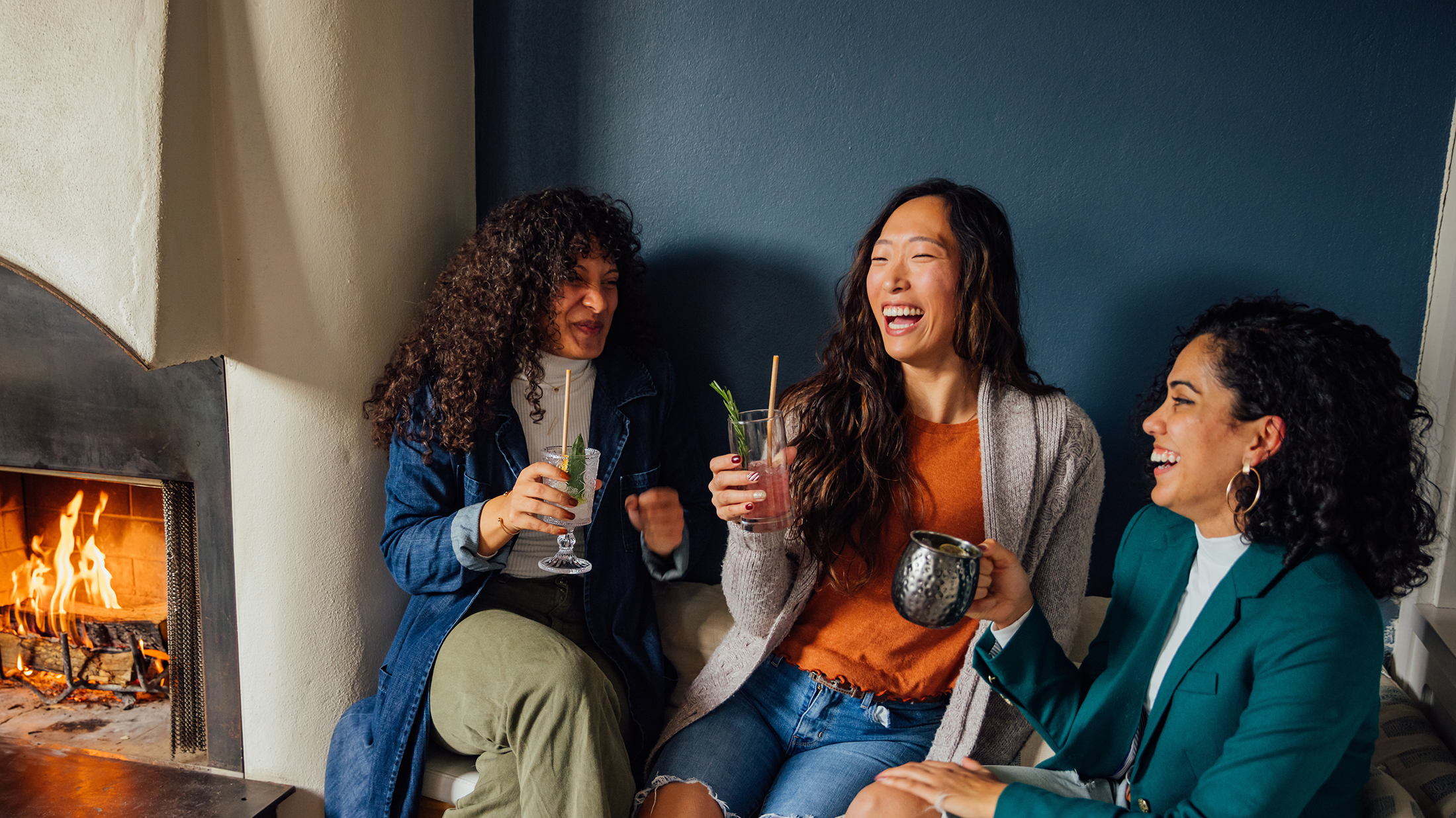 Trois amis rient ensemble tout en dégustant des cocktails près d'une cheminée crépitante au Suraya.
