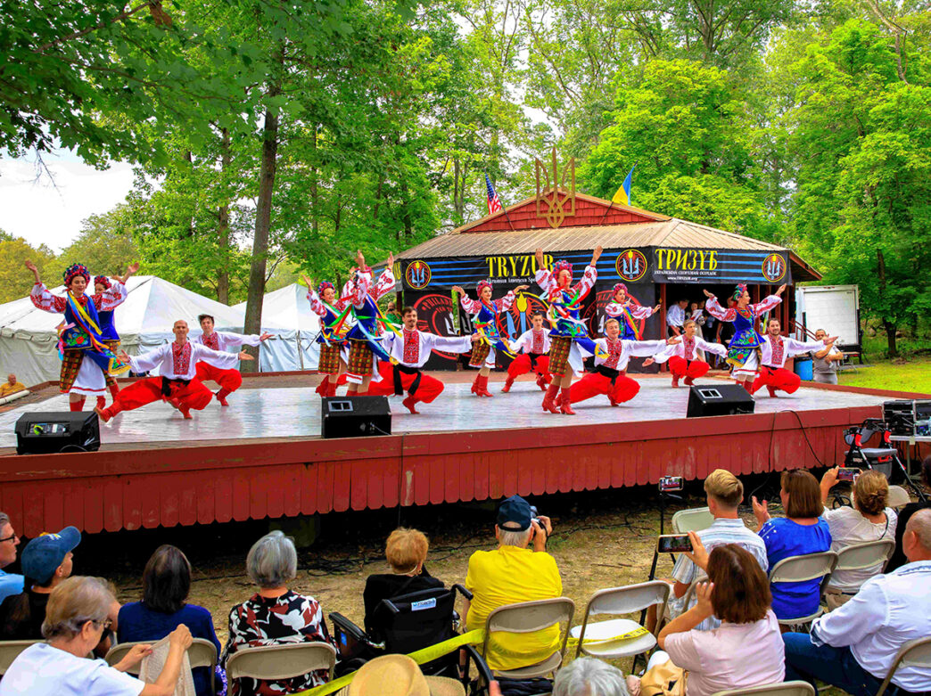 Performers in vibrant traditional Ukrainian attire dance on an outdoor stage surrounded by tall green trees and an audience sitting in folding chairs.