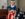 A Betsy Ross impersonator in colonial-era clothing sits indoors hand-sewing a large American flag. Wearing a blue dress, white apron, and headscarf, she carefully stitches red-and-white stripes while a blue field with white stars rests on a nearby table. Period furniture, a candle, and curtained windows create a historic 18th-century setting.