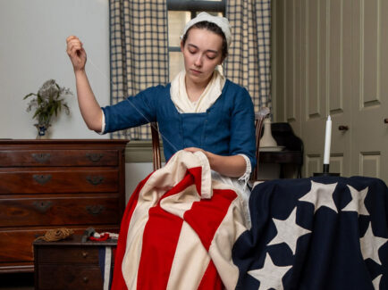 A Betsy Ross impersonator in colonial-era clothing sits indoors hand-sewing a large American flag. Wearing a blue dress, white apron, and headscarf, she carefully stitches red-and-white stripes while a blue field with white stars rests on a nearby table. Period furniture, a candle, and curtained windows create a historic 18th-century setting.