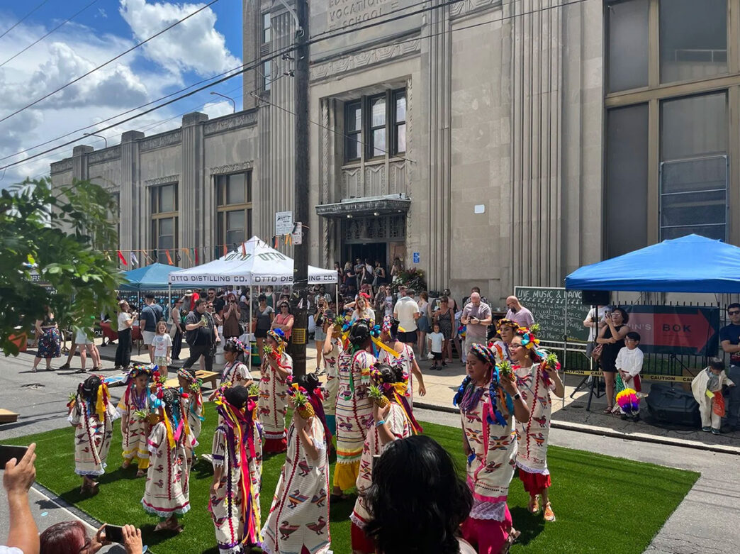 Dancers in colorful traditional attire perform on a grassy plaza during a b[l]ok party at Bok in South Philadelphia. Spectators gather around the outdoor performance space, while vendors, live music, and activities fill the street in front of the historic Bok building on a sunny day.