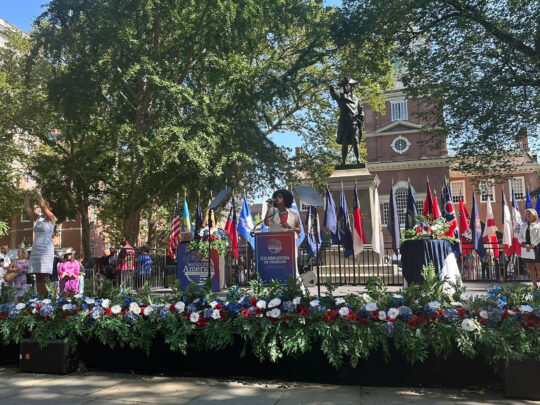 A singer stands on an outdoor stage in front of Independence Hall, where patriotic floral displays and military flags decorate the stage during the Celebration of Freedom Ceremony.