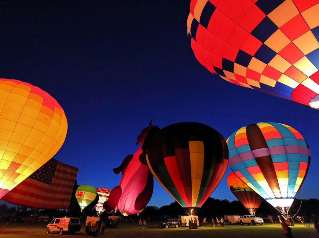 Colorful hot air balloons glow against the night sky during the Chester County Hot Air Balloon Festival. Several illuminated balloons in bright patterns and shapes are tethered on a grassy field, while spectators, vehicles, and an American flag are visible below during an evening balloon glow event.