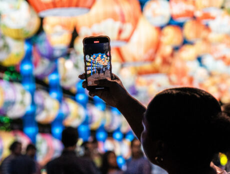 A visitor holds up a smartphone to photograph rows of colorful illuminated lanterns at Philadelphia’s Chinese Lantern Festival, with glowing lantern displays filling the background and festivalgoers blurred below.