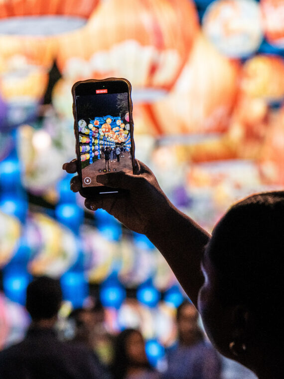 A visitor holds up a smartphone to photograph rows of colorful illuminated lanterns at Philadelphia’s Chinese Lantern Festival, with glowing lantern displays filling the background and festivalgoers blurred below.