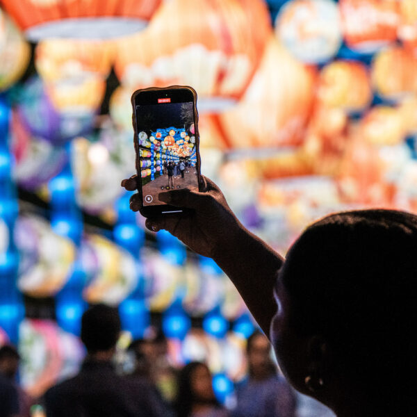 Un visiteur brandit son smartphone pour photographier des rangées de lanternes colorées lors du festival des lanternes chinoises de Philadelphie, avec des lanternes lumineuses en arrière-plan et des festivaliers en arrière-plan.