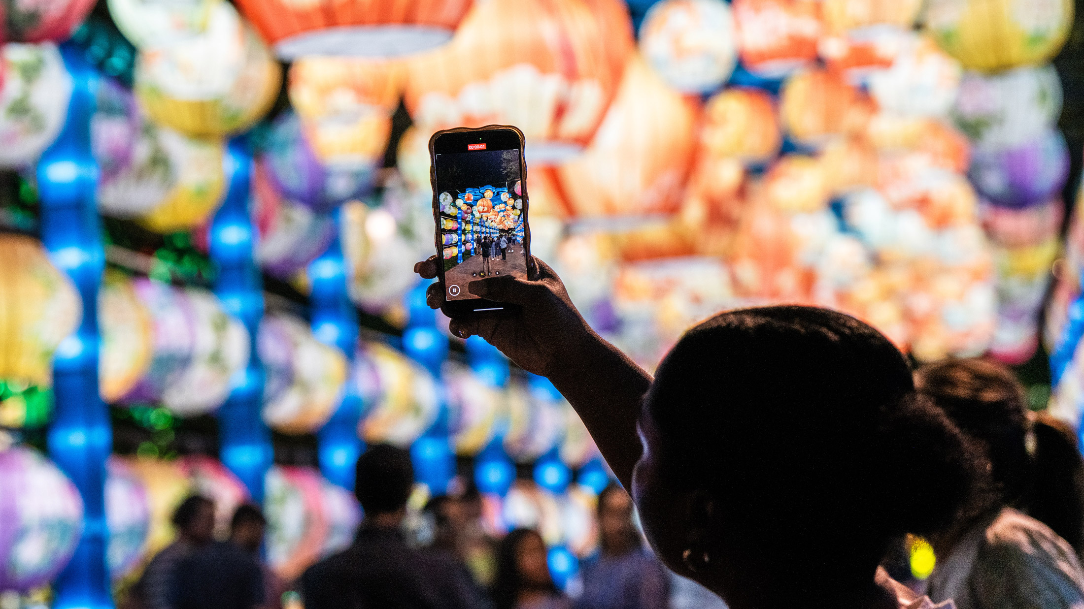 Um visitante segura um smartphone para fotografar fileiras de lanternas coloridas e iluminadas no Festival de Lanternas Chinesas da Filadélfia, com exibições de lanternas brilhantes preenchendo o fundo e os frequentadores do festival desfocados abaixo.