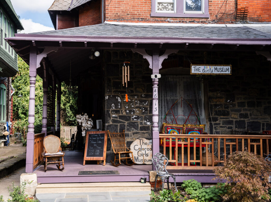 The Colored Girls Museum, a historic rowhouse in Philadelphia, features a welcoming front porch with rocking chairs, colorful decor, and a museum sign above the entrance. Hand-painted signs and artwork highlight the museum’s community-centered atmosphere and celebration of Black women’s stories and culture.