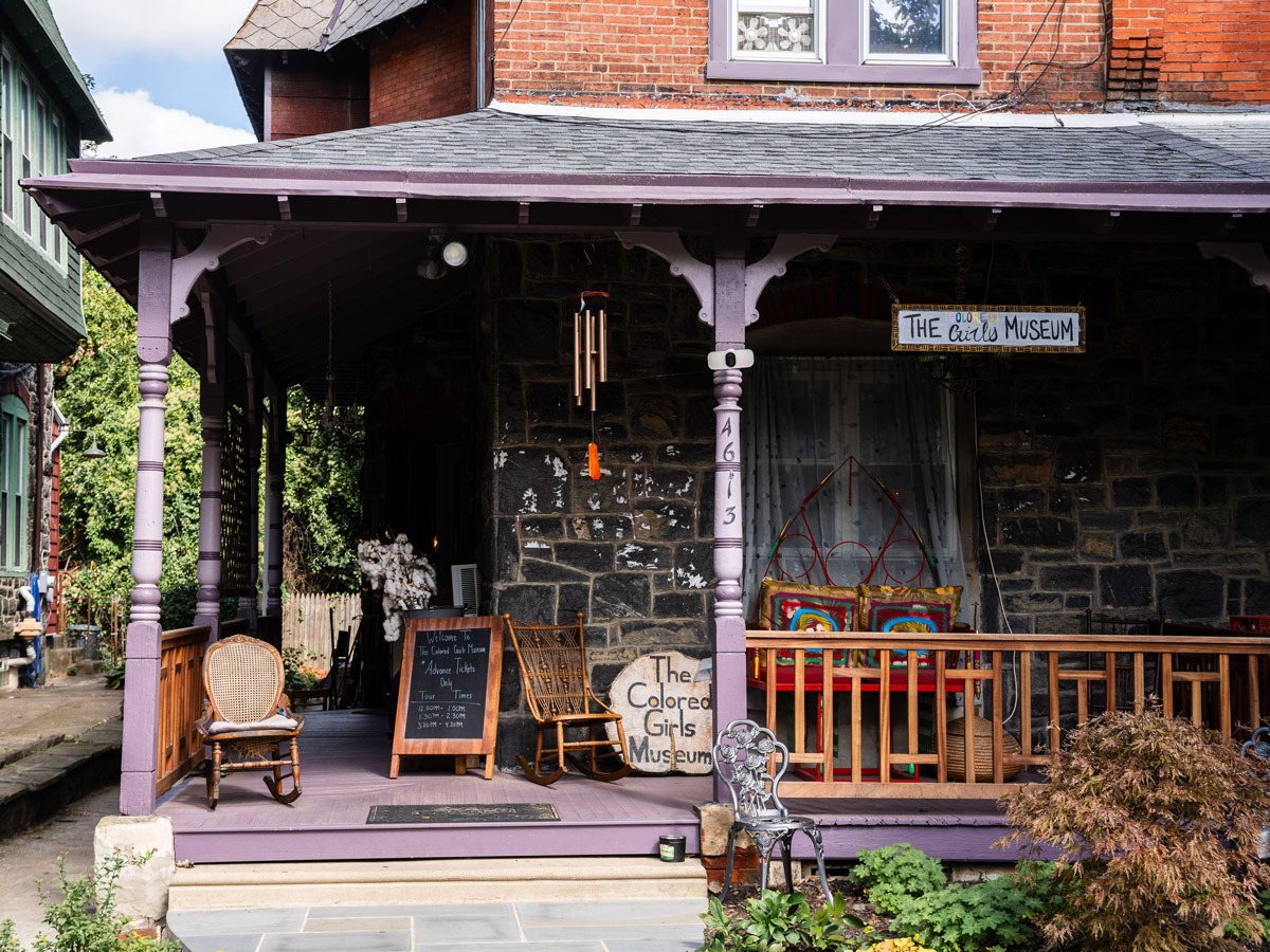 The Colored Girls Museum, a historic rowhouse in Philadelphia, features a welcoming front porch with rocking chairs, colorful decor, and a museum sign above the entrance. Hand-painted signs and artwork highlight the museum’s community-centered atmosphere and celebration of Black women’s stories and culture.