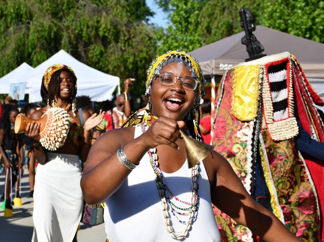 Participants in Delaware County’s Annual Juneteenth Celebration parade along a sunny outdoor walkway wearing colorful African-inspired attire and beaded accessories. In the foreground, a smiling participant rings a hand bell while others play percussion instruments and dance, creating a vibrant scene that celebrates Black culture, community, and freedom.