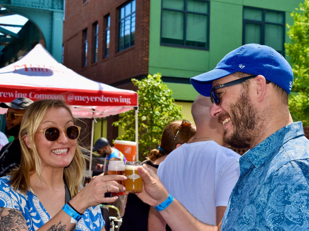 Two attendees smile and toast with small glasses of beer at Evil Genius Brewing Company’s Fishtown Beer Festival. Standing outdoors among fellow festivalgoers, the pair wear sunglasses and casual summer clothing as they sample craft beer near brewery tents and vendors in Philadelphia’s Fishtown neighborhood.