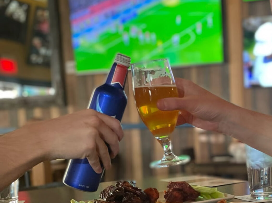 Two people clink a bottled beer and a pint glass of golden beer over a plate of chicken wings at Fadó Irish Pub. A large television in the background displays a soccer match, creating a lively sports bar atmosphere.