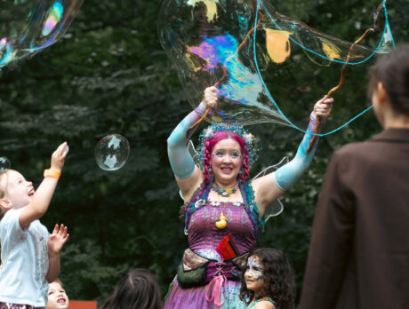 A costumed fairy performer with bright pink hair and iridescent wings creates giant shimmering bubbles amid a crowd ofchildren during Fairy Fest at the Schuylkill Center for Environmental Education.