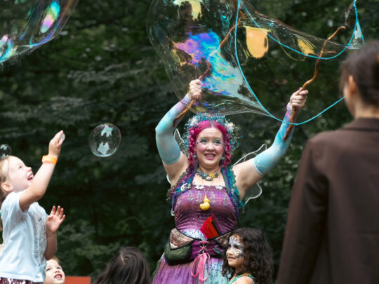 Une fée costumée aux cheveux rose vif et aux ailes irisées crée des bulles géantes et scintillantes au milieu d'une foule d'enfants lors de la fête des fées au centre Schuylkill pour l'éducation à l'environnement.