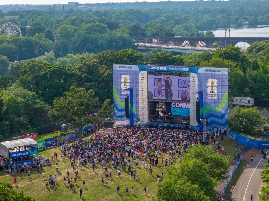 An aerial view shows attendees gathered at Philadelphia’s FIFA Fan Festival around a large stage and video screen. Set within a green park landscape near the Delaware River, the event features live entertainment, fan activities, and FIFA World Cup 26 branding throughout the festival grounds.