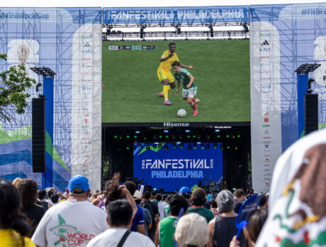 Crowds gather at Philadelphia’s FIFA Fan Festival to watch a live soccer match on a giant video screen. The outdoor event features a large stage branded for FIFA World Cup 26 and FIFA Fan Festival Philadelphia, with fans in team jerseys and national colors watching the action together in a festive atmosphere.