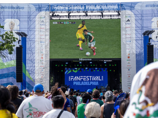 La foule se rassemble au FIFA Fan Festival de Philadelphie pour regarder un match de football en direct sur un écran vidéo géant. L'événement en plein air comprend une grande scène portant la marque de la Coupe du Monde de la FIFA 26 et du FIFA Fan Festival Philadelphia, avec des supporters vêtus de maillots d'équipes et de couleurs nationales qui regardent l'action ensemble dans une atmosphère festive.