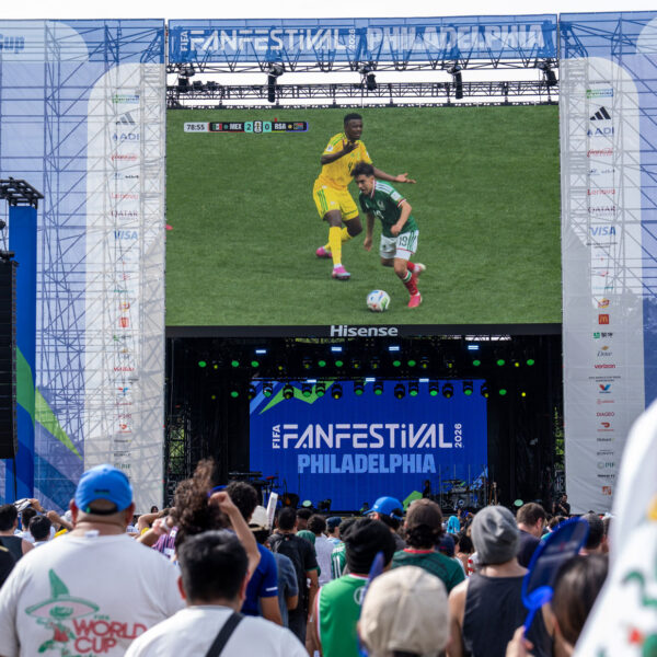 La foule se rassemble au FIFA Fan Festival de Philadelphie pour regarder un match de football en direct sur un écran vidéo géant. L'événement en plein air comprend une grande scène portant la marque de la Coupe du Monde de la FIFA 26 et du FIFA Fan Festival Philadelphia, avec des supporters vêtus de maillots d'équipes et de couleurs nationales qui regardent l'action ensemble dans une atmosphère festive.