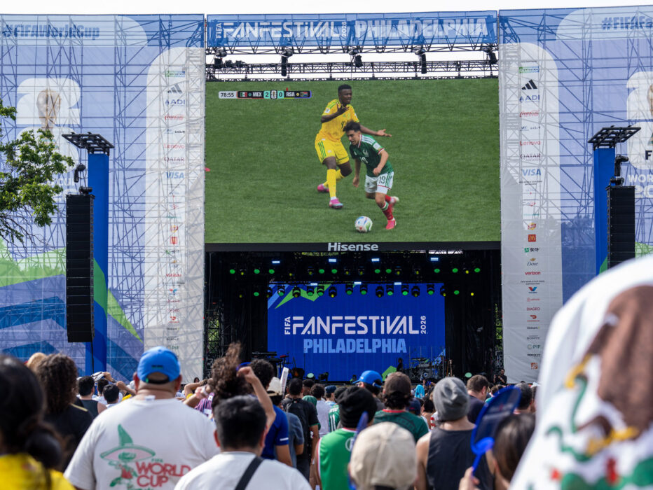 Multidões se reúnem no FIFA Fan Festival da Filadélfia para assistir a uma partida de futebol ao vivo em uma tela de vídeo gigante. O evento ao ar livre apresenta um grande palco com a marca da Copa do Mundo da FIFA 26 e do FIFA Fan Festival Philadelphia, com torcedores vestidos com camisas de times e cores nacionais assistindo à ação juntos em uma atmosfera festiva.