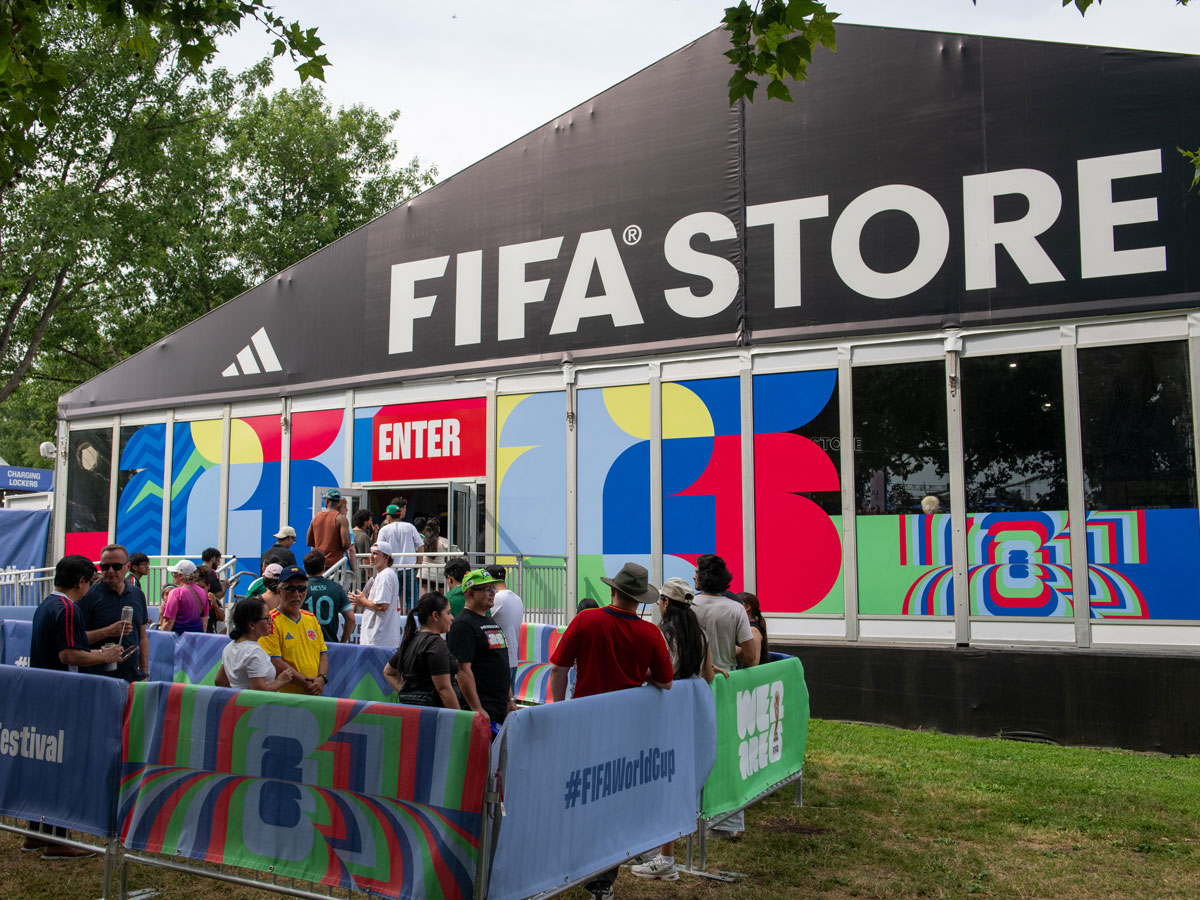 Los visitantes hacen fila frente a la tienda de la FIFA en el Festival de Aficionados de la FIFA de Filadelfia, donde se exhiben coloridos elementos promocionales de la Copa Mundial de la FIFA 26 en el escaparate y en las barreras que separan a la multitud.