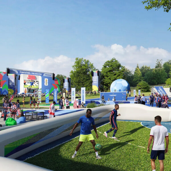 Visitors play a casual soccer game on a small field at the FIFA World Cup 2026 Fan Festival in Philadelphia, with crowds, interactive activities, and a large viewing stage visible in the background.