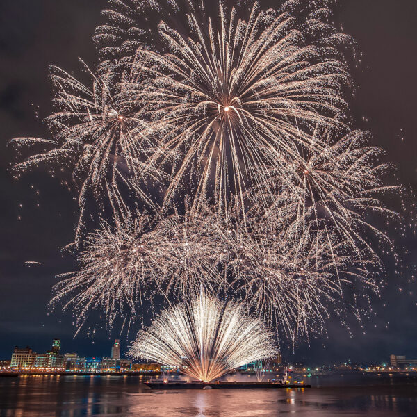 Fogos de artifício brancos e brilhantes explodiram acima do Delaware River Waterfront durante uma comemoração do Dia da Independência, iluminando o céu noturno e refletindo na água, com o horizonte da Filadélfia ao fundo.