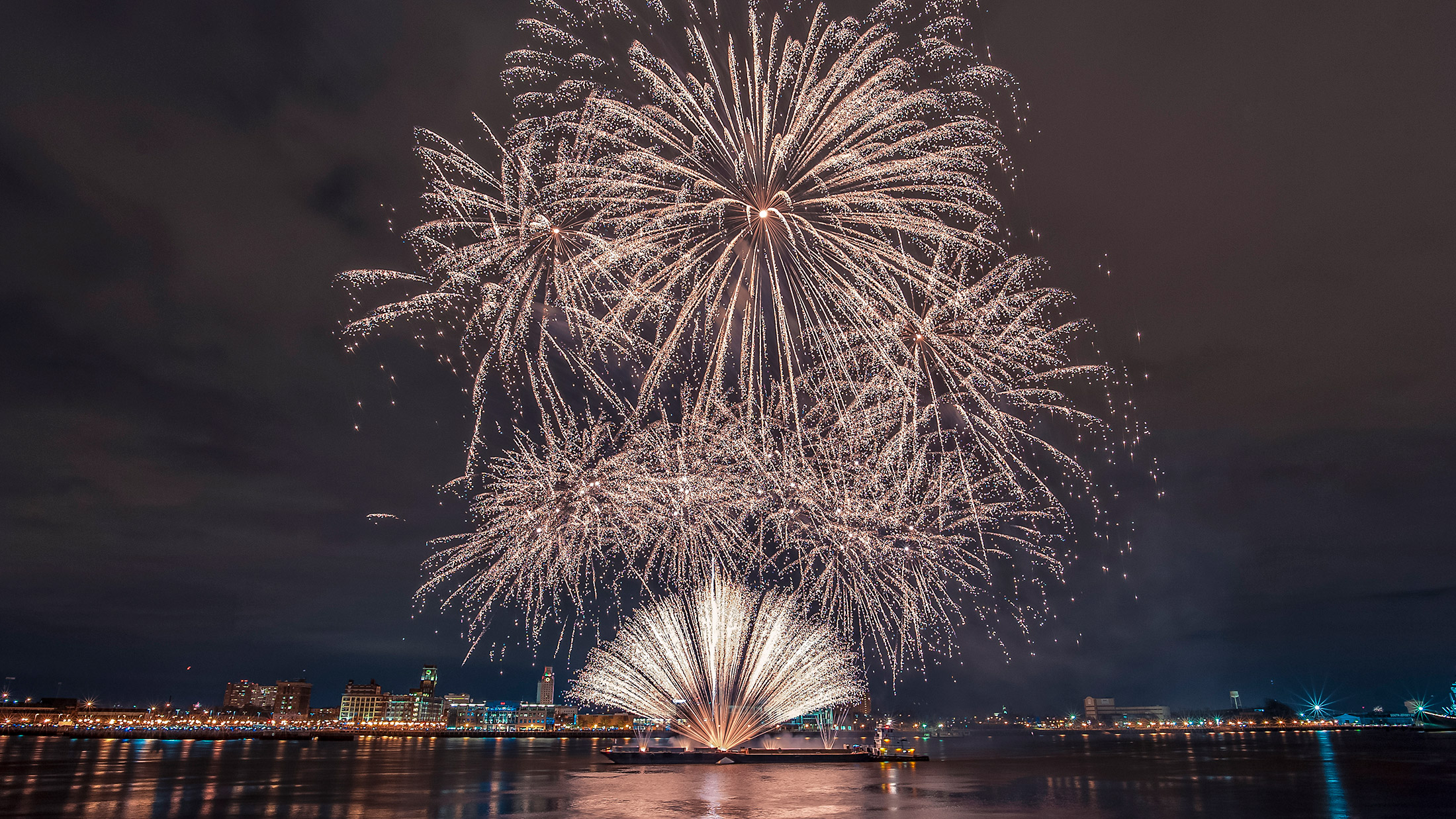 Bright white fireworks burst above the Delaware River Waterfront during an Independence Day celebration, illuminating the night sky and reflecting on the water with the Philadelphia skyline in the background.
