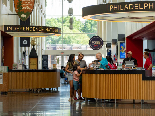Visitors gather at information counters inside Philadelphia’s Independence Visitor Center, where staff assist guests with trip planning and tickets. Bright signage for Independence National Historical Park and local attractions hangs above the spacious, sunlit lobby with floor-to-ceiling windows.