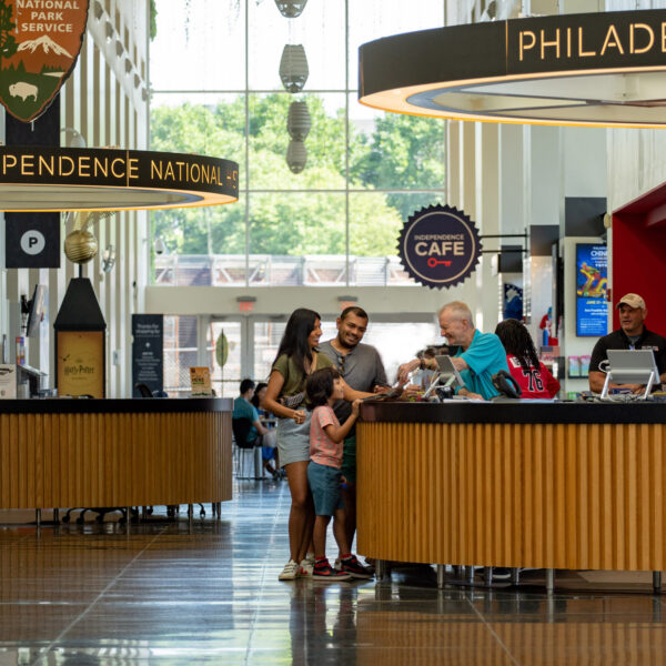 Les visiteurs se rassemblent aux comptoirs d'information du Independence Visitor Center de Philadelphie, où le personnel les aide à planifier leur voyage et à acheter leurs billets. Des panneaux lumineux indiquant le parc historique national de l'Indépendance et les attractions locales sont suspendus au-dessus du hall d'entrée spacieux et ensoleillé, doté de fenêtres allant du sol au plafond.
