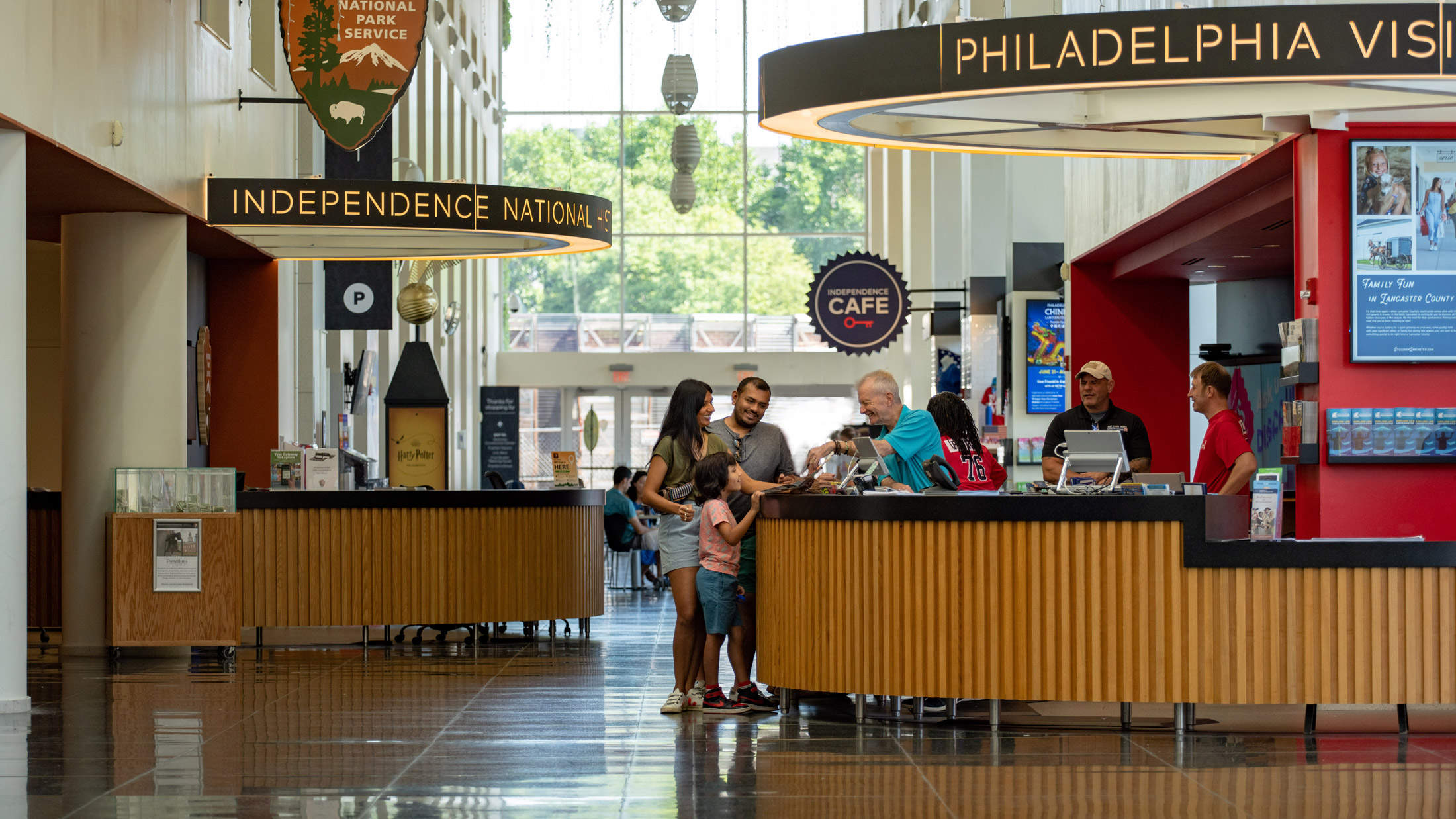 Visitors gather at information counters inside Philadelphia’s Independence Visitor Center, where staff assist guests with trip planning and tickets. Bright signage for Independence National Historical Park and local attractions hangs above the spacious, sunlit lobby with floor-to-ceiling windows.