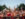 A packed crowd cheers and raises their hands during Philadelphia’s Fourth of July concert on the Benjamin Franklin Parkway. Concertgoers stand behind barricades, many wearing patriotic red, white, and blue clothing, with the Center City skyline visible above the tree-lined boulevard at dusk.