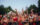 A packed crowd cheers and raises their hands during Philadelphia’s Fourth of July concert on the Benjamin Franklin Parkway. Concertgoers stand behind barricades, many wearing patriotic red, white, and blue clothing, with the Center City skyline visible above the tree-lined boulevard at dusk.