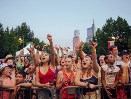 A packed crowd cheers and raises their hands during Philadelphia’s Fourth of July concert on the Benjamin Franklin Parkway. Concertgoers stand behind barricades, many wearing patriotic red, white, and blue clothing, with the Center City skyline visible above the tree-lined boulevard at dusk.