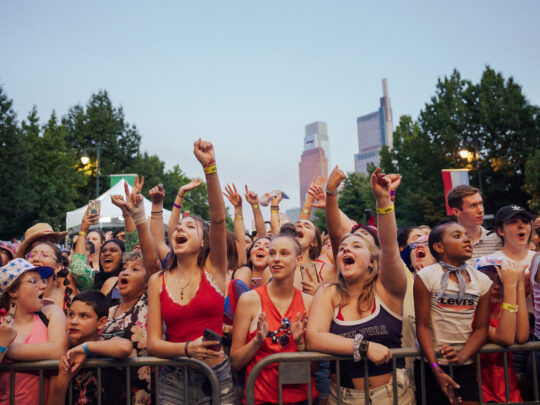 A packed crowd cheers and raises their hands during Philadelphia’s Fourth of July concert on the Benjamin Franklin Parkway. Concertgoers stand behind barricades, many wearing patriotic red, white, and blue clothing, with the Center City skyline visible above the tree-lined boulevard at dusk.