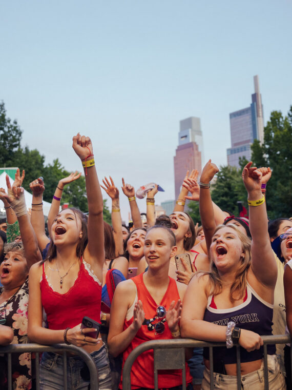 Uma multidão lotada aplaude e levanta as mãos durante o show de 4 de julho na Filadélfia, no Benjamin Franklin Parkway. Os frequentadores do show ficam atrás de barricadas, muitos vestindo roupas patrióticas vermelhas, brancas e azuis, com o horizonte do centro da cidade visível acima da avenida arborizada ao anoitecer.