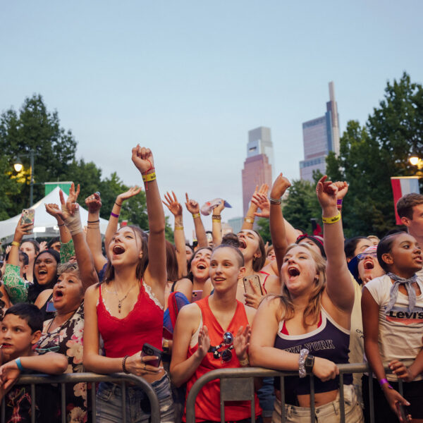 Uma multidão lotada aplaude e levanta as mãos durante o show de 4 de julho na Filadélfia, no Benjamin Franklin Parkway. Os frequentadores do show ficam atrás de barricadas, muitos vestindo roupas patrióticas vermelhas, brancas e azuis, com o horizonte do centro da cidade visível acima da avenida arborizada ao anoitecer.
