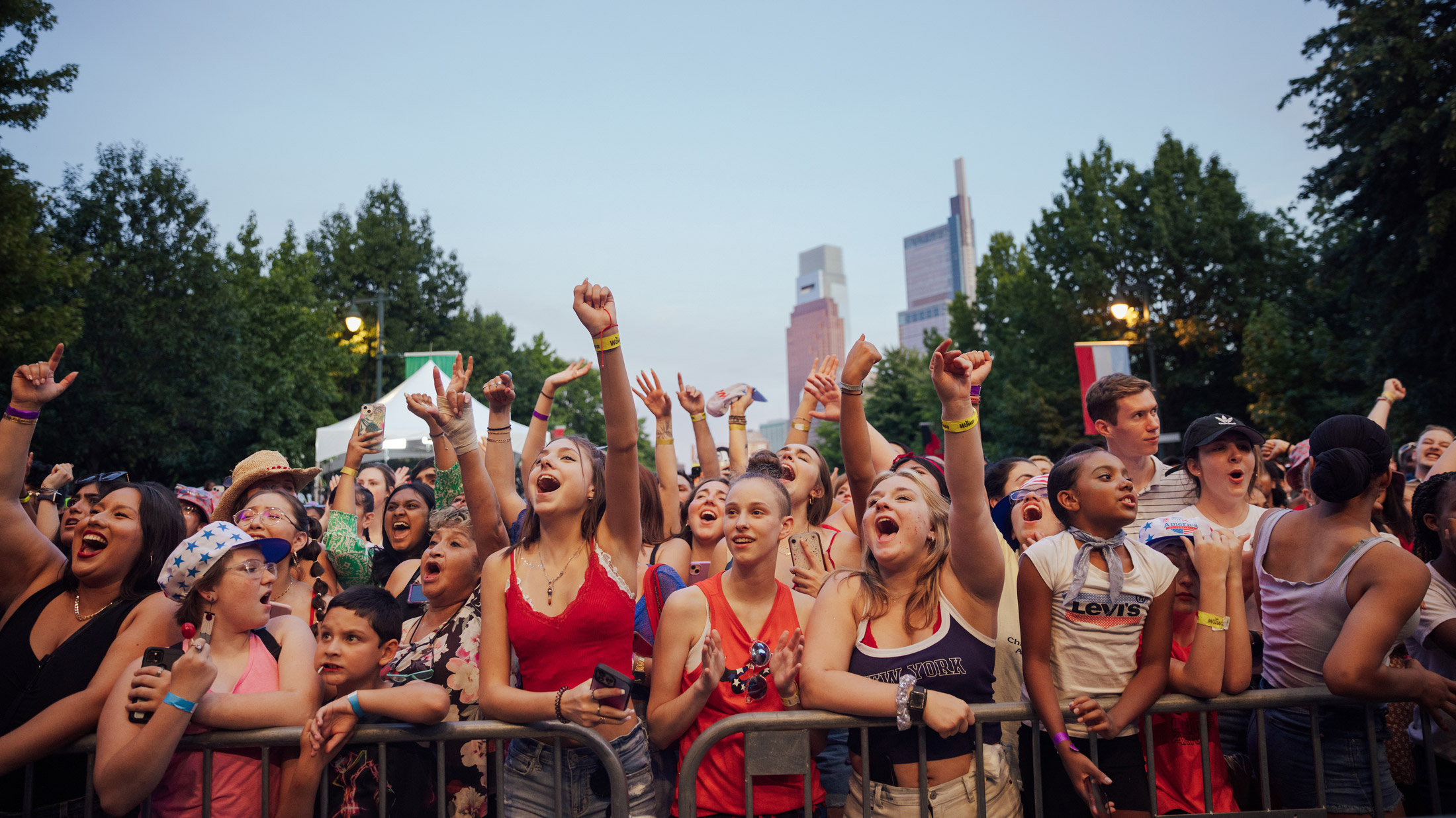 A packed crowd cheers and raises their hands during Philadelphia’s Fourth of July concert on the Benjamin Franklin Parkway. Concertgoers stand behind barricades, many wearing patriotic red, white, and blue clothing, with the Center City skyline visible above the tree-lined boulevard at dusk.